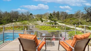 A poolside area with wicker chairs and a table, overlooking a landscaped garden and a pool. - Dunes Summer Retreat • Easy Walk to Private Ocean Beach (Amagansett)
