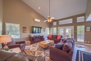 A spacious living room with a large sofa, a coffee table, and a TV. There are multiple windows and a ceiling fan.
