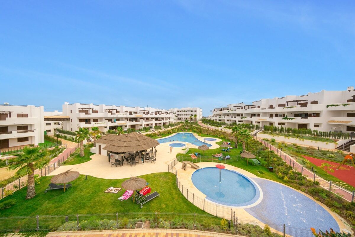 Ein Resort mit mehreren weißen Gebäuden, einem zentralen Schwimmbecken und einem angelegten Gartenbereich mit Palmen und Liegestühlen.