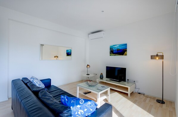 A modern living room with a black sofa, a glass coffee table, a flat-screen TV, a floor lamp, and artwork on the walls.