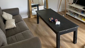 Een woonkamer met een grijze bank, een zwarte salontafel, een vloerlamp, een tv op een dressoir en een boekenkast.