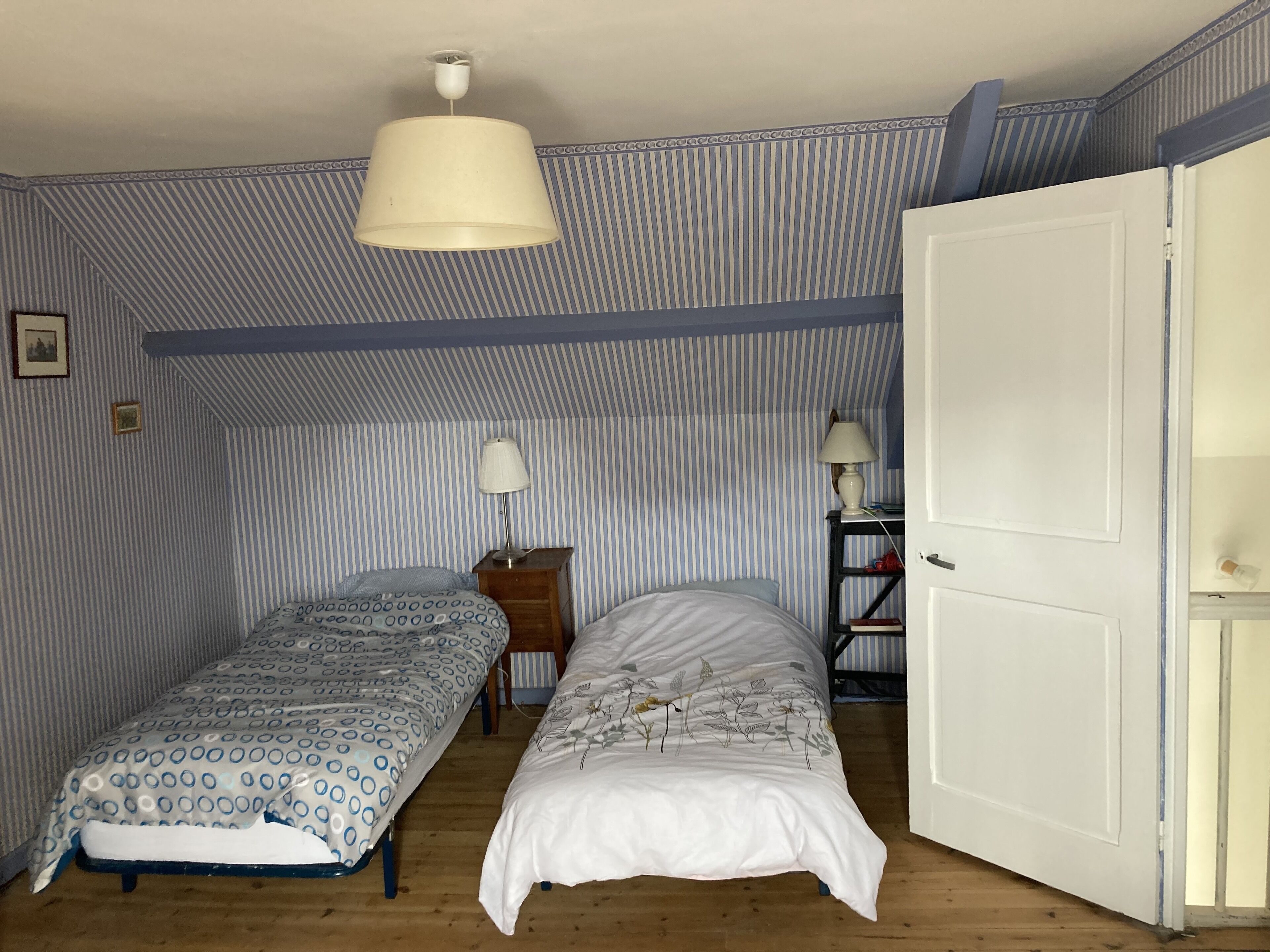 A room with two single beds, a wooden floor, striped wallpaper, a white door, and a hanging lamp.