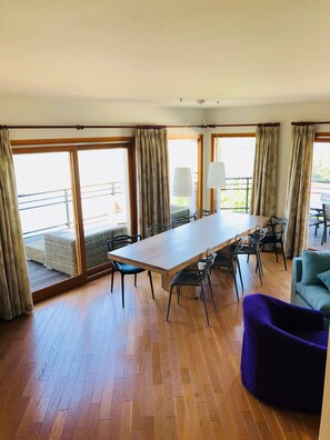 A spacious room with a large wooden table, multiple chairs, a sofa, and large windows with curtains.