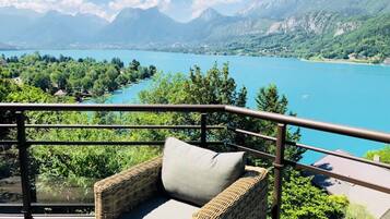Une chaise en osier avec un coussin, placée sur un balcon donnant sur un lac, avec des montagnes en arrière-plan.