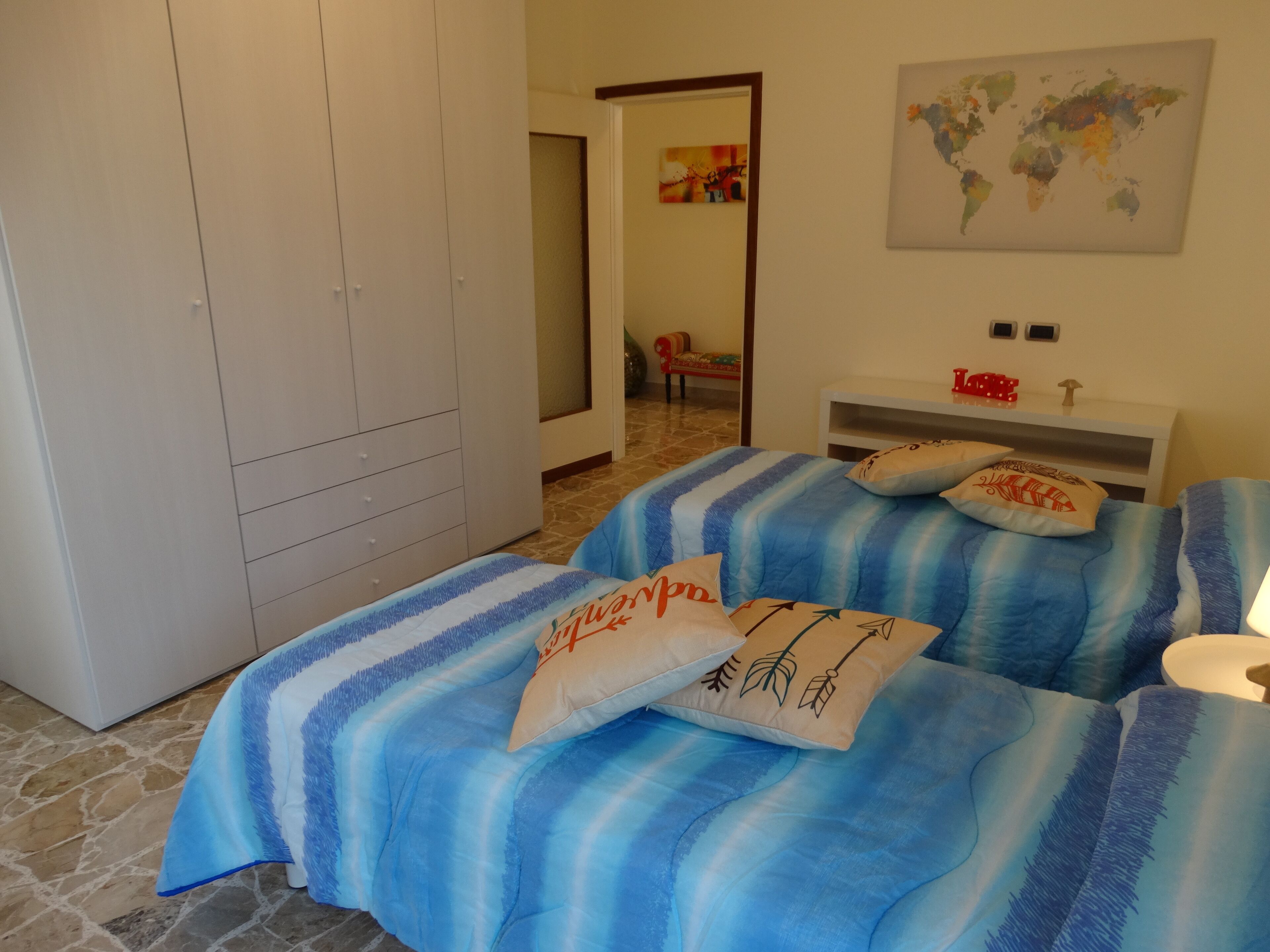 A bedroom with a bed covered in blue and white striped bedding, a white wardrobe, a world map on the wall, and a small table with decorative items.