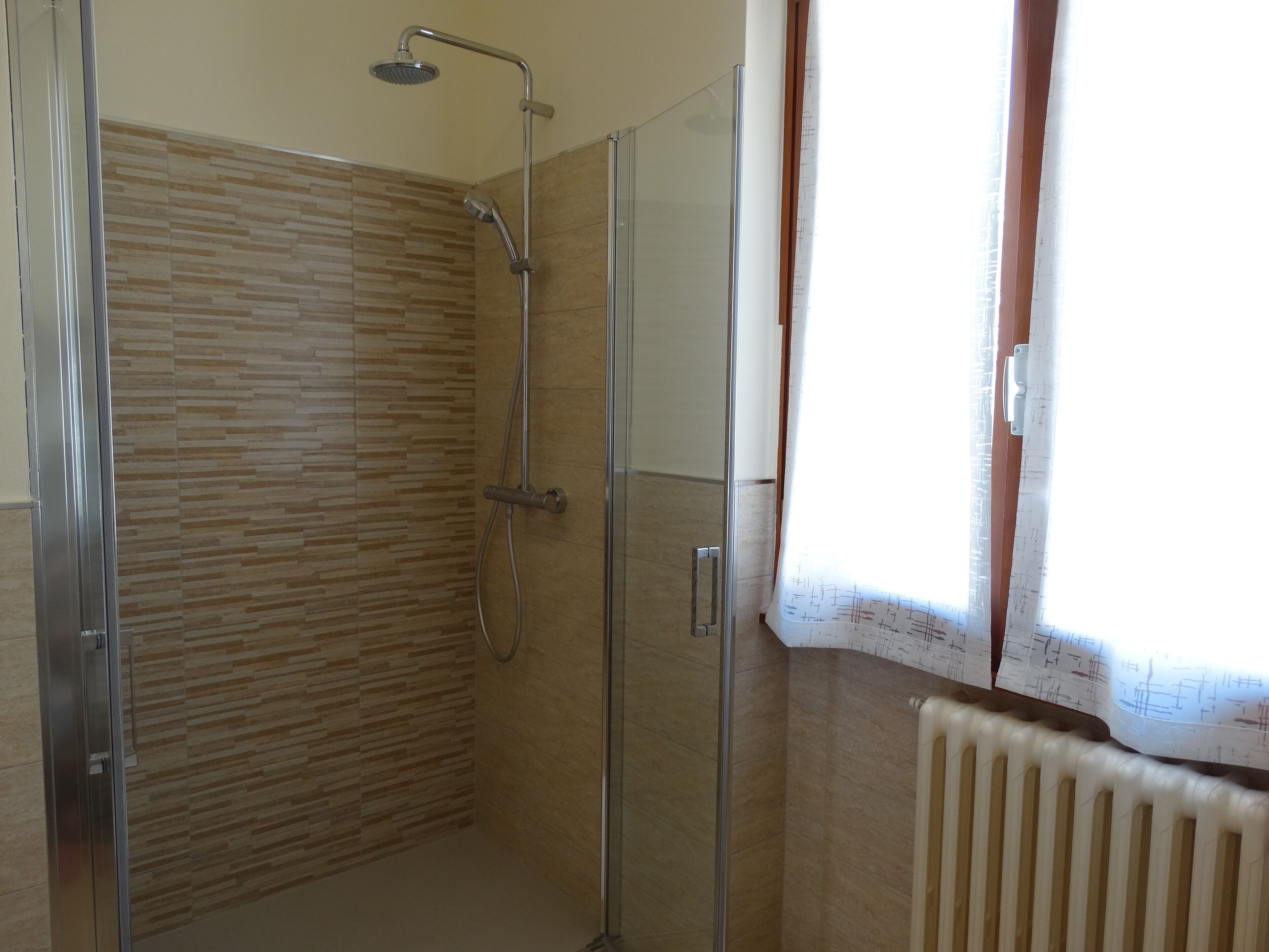 A bathroom with a shower, a radiator, and a window with curtains.