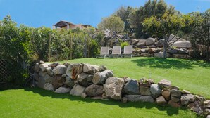 Un muro de piedra con dos sillones de descanso en un área cubierta de césped, rodeado de árboles y una casa al fondo.
