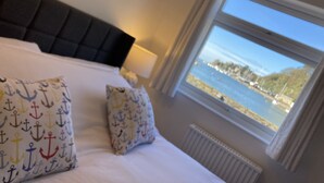 A bedroom with a large bed, two pillows with anchor patterns, a dark headboard, a window with a view of a body of water and boats, and a white radiator. - Sea View Holiday Cottage Porthmadog (Porthmadog)