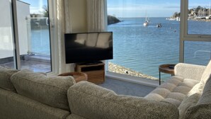 A living room with a large TV, beige sofas, and a view of the sea with boats. - Sea View Holiday Cottage Porthmadog (Porthmadog)