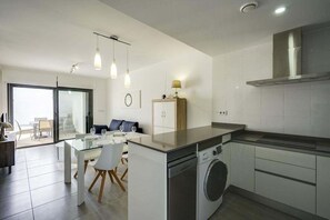 A modern kitchen with a dishwasher, a built-in oven, and a stainless steel range hood. There is a dining area with a glass table and chairs, and a sofa in the background. - Apartamento Tulipanes (Pilar de la Horadada) (Pilar de la Horadada)