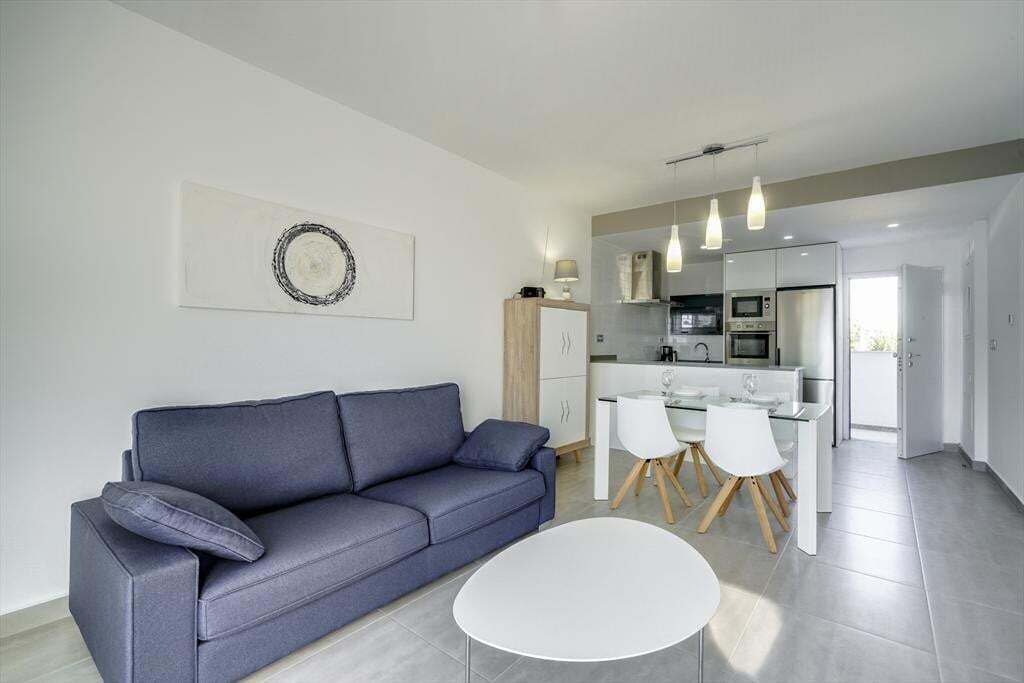 Ein modernes Wohnzimmer mit grauem Sofa, einem kleinen runden Tisch und einem Essbereich mit weißen Stühlen und einem Glastisch.