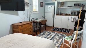 Una habitación funcional con cama, cómoda de madera, mesa de comedor, sillas y una cocina equipada con microondas, refrigerador y escoba.