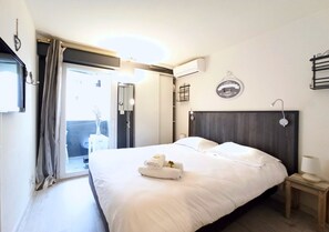 A hotel room with a bed, a bedside table, a nightstand, a window with curtains, and an air conditioning unit. - Viva Riviera Residence Palazzio 1 Bedroom  Bellevue (Cannes)