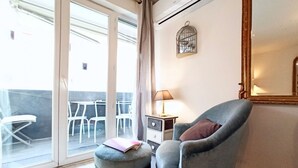 Una habitación con un amplio ventanal corredero que da a un espacio exterior con sillas y mesas blancas. Hay un sillón gris con cojín, un pequeño banquillo con un libro y una mesita de noche con una lámpara.