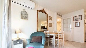 Una habitación con un sillón azul, una mesita con una lámpara, una mesa de comedor con sillas, un espejo grande y una jaula de pájaros en la pared.