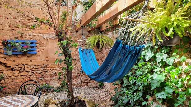 Un espacio de estar al aire libre con aspecto rústico, que incluye una hamaca, una mesita y plantas en macetas frente a un muro de piedra.