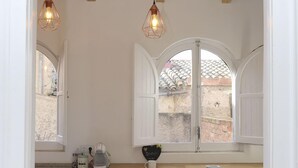 Una cocina con techo de vigas de madera, dos lámparas colgantes, un ventanal con contraventanas blancas, una encimera con un hervidor y un cuenco, y dos taburetes.