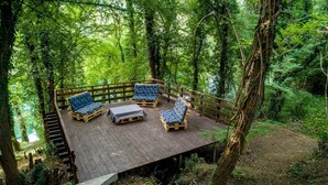 A wooden deck with four blue cushioned chairs and a table, surrounded by dense forest. - Luxury house with private pool, sauna, hot tub and river on the property (Danilovgrad)