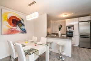 A modern kitchen with stainless steel appliances, a dining area with a glass table, and a colorful abstract painting on the wall. - Apartamento Begonia (Villamartin) (Orihuela Costa)