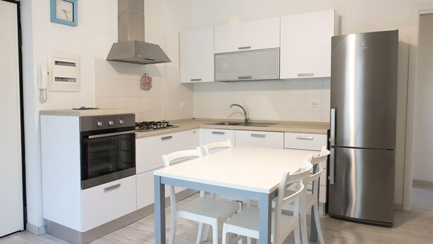 A modern kitchen with white cabinets, a stainless steel refrigerator, a gas stove, and a white dining table with chairs. - S027 - Sirolo, new three-room apartment with terrace in the center (Sirolo)