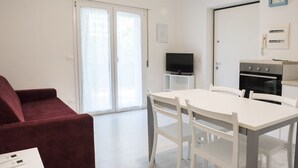 A modern living room with a red sofa, a white dining table with chairs, a flat-screen TV on a white stand, and a kitchen area with an oven. - S027 - Sirolo, new three-room apartment with terrace in the center (Sirolo)