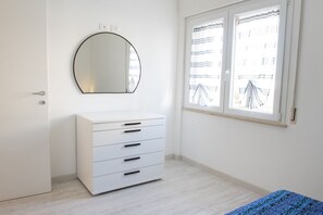 Ett sovrum med en vit byrå, en rund spegel, ett fönster med gardiner och en säng med ett blått, mönstrat överkast.