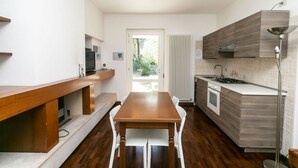 A modern kitchen with a wooden dining table, white chairs, a built-in oven, and a window overlooking a garden.