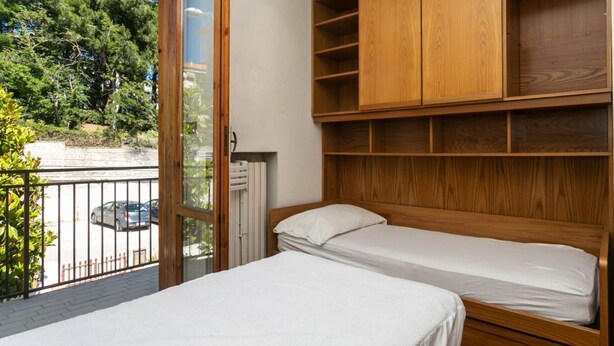 A bedroom with two beds, wooden shelving, and a view of trees and a parking area.