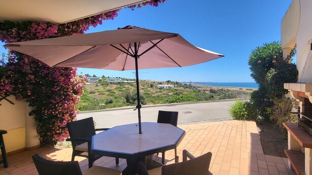 Une terrasse extérieure avec une table et des chaises, un grand parasol et une vue sur la mer et le paysage environnant.