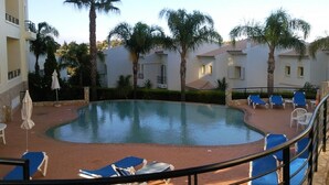 Une piscine entourée de palmiers et d’un bâtiment doté de balcons, avec des chaises longues bleues et des parasols.