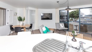 A modern living room with a dining area, a large window with a view, a flat-screen TV, and a kitchen with a sink. - Spacious Large Modern Apartment in Fantastic Location (Tauranga)