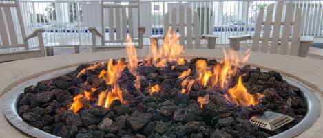 A circular fire pit with burning charcoal in a covered outdoor area with white chairs and a view of a parking lot.