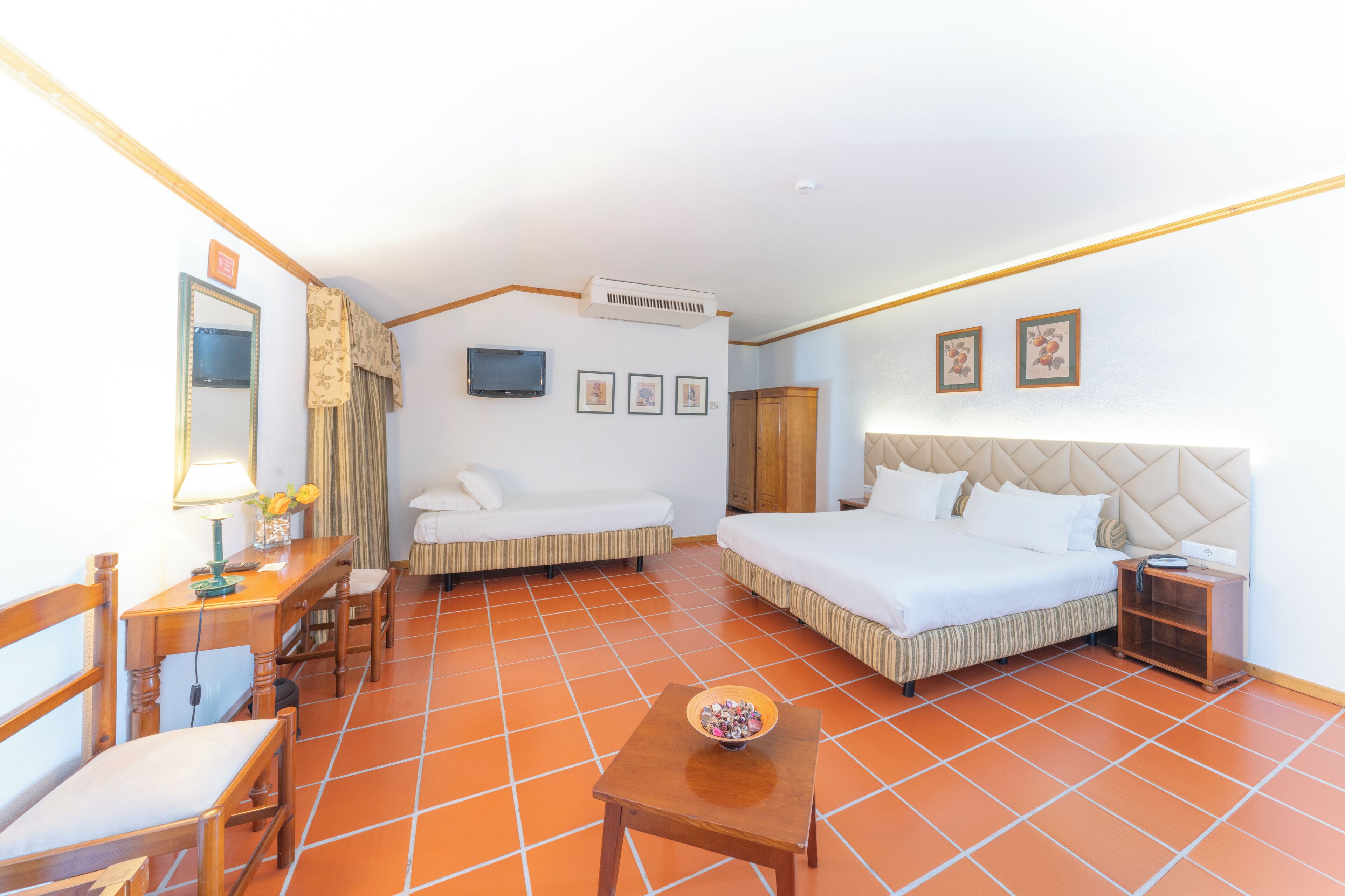 A hotel room with a bed, a sofa, a desk with a lamp, a small table with a bowl, and a TV on the wall.