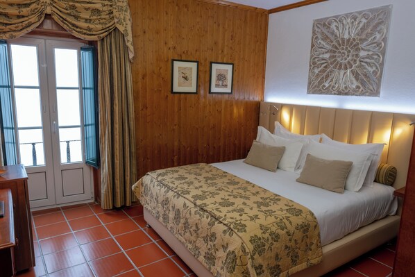 A bedroom with a wooden paneled wall, a bed with a floral bedspread, two framed pictures, and a large decorative piece above the bed. - Hotel Dom Manuel (Marvao)