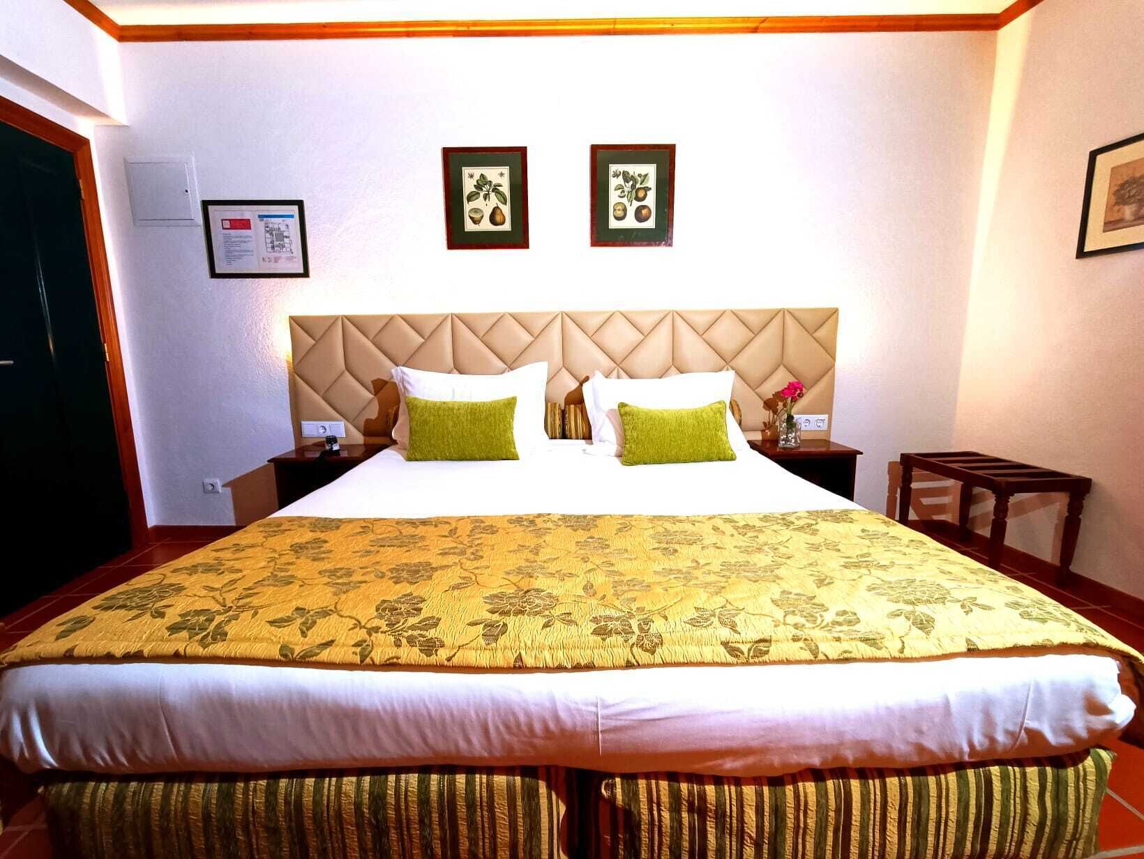 Una habitación de hotel con una cama grande, cabecera con diseño de diamantes, dos cuadros en la pared y una pequeña mesa con un jarrón de flores.