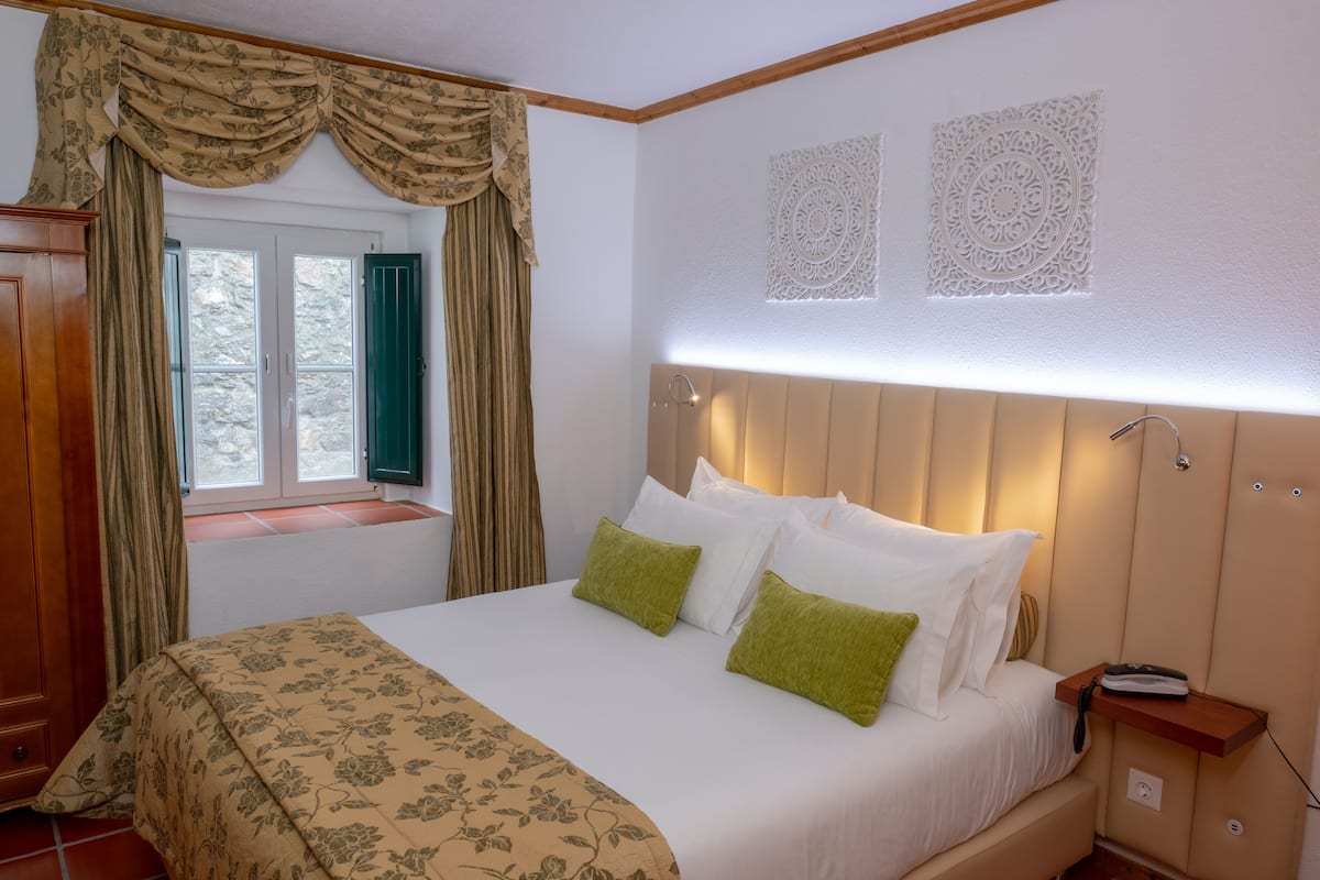 Ein ordentlich bezogenes Bett mit einer blumigen Tagesdecke, ein Nachttisch mit Lampe und ein Fenster mit Vorhängen.
