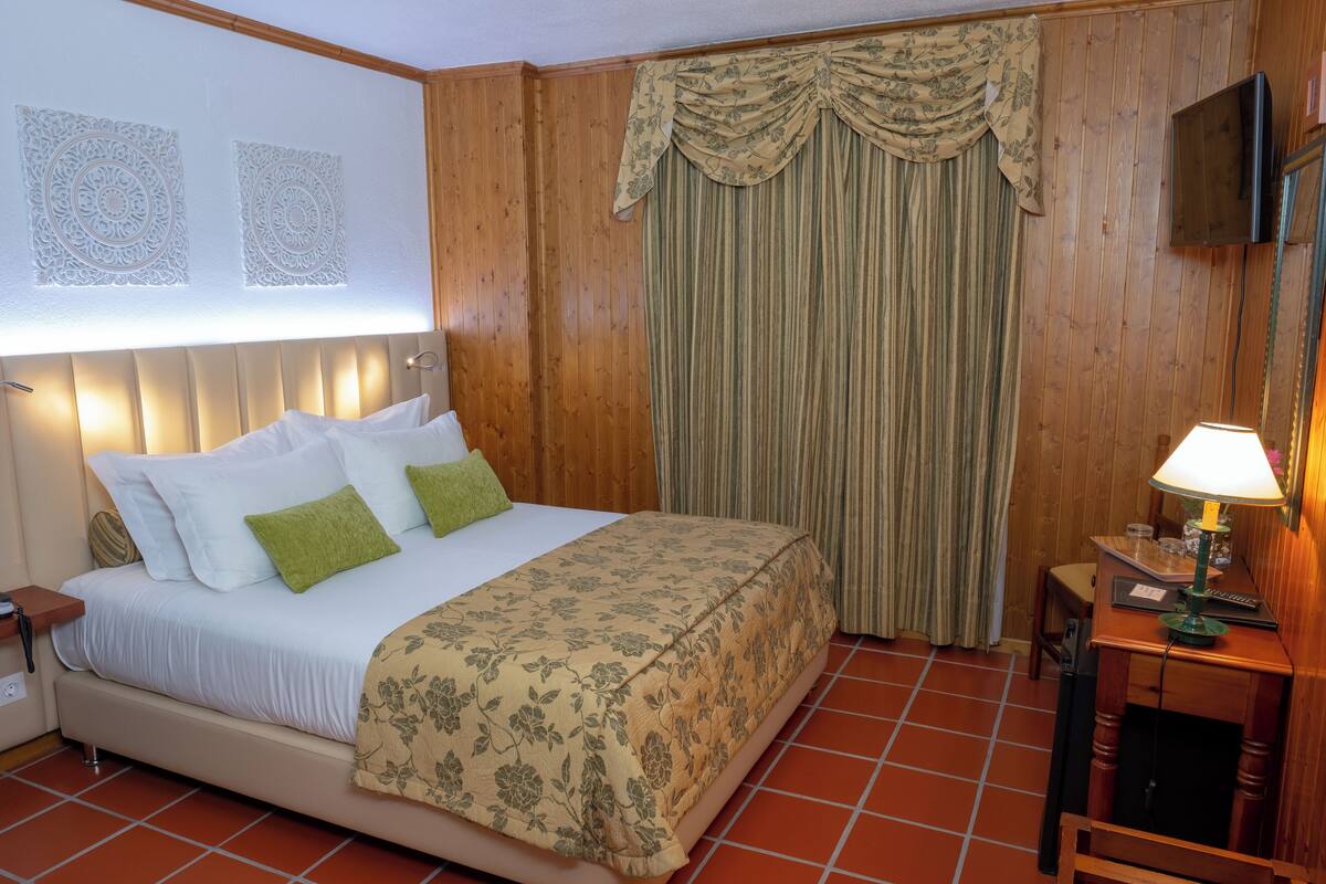 Ein ordentlich eingerichtetes Hotelzimmer mit Bett, Nachttisch mit Lampe, einem an der Wand montierten Fernseher und einem Vorhang.