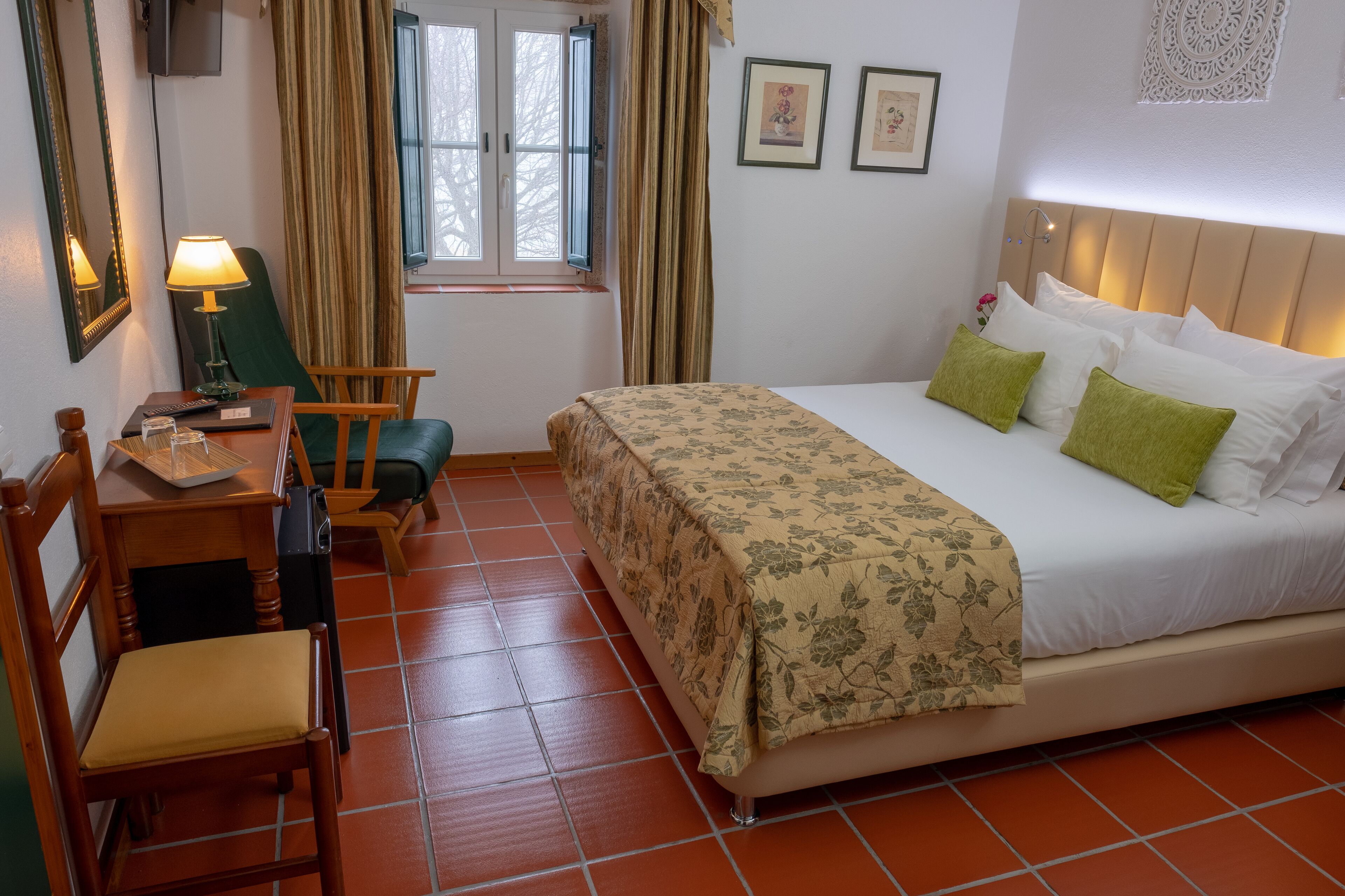A bedroom with a bed, a desk with a lamp, a chair, and a window with curtains.