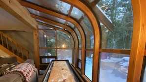 Ein gemütliches Zimmer mit Shuffleboard-Tisch, Holzwänden und großen Fenstern mit Blick auf die verschneite Landschaft.