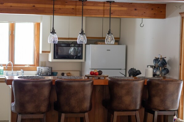 A kitchen with wooden ceiling beams, a white refrigerator, a microwave, and a bar area with four leather chairs.