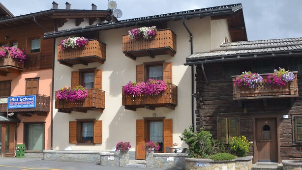 A building with wooden balconies adorned with flowers, a sign for a ski school, and a parking area. - Ringemel-2 apartments (Livigno)