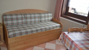 A wooden sofa with plaid cushions in a room with a window, a radiator, and a table with a checkered tablecloth. - Ringemel-2 apartments (Livigno)