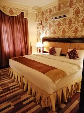 A hotel room with a large bed, a headboard, a wall-mounted air conditioner, and a patterned wall. - Dmas Hotel (Muscat)