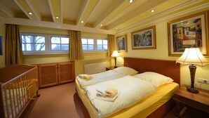 Une chambre d’hôtel avec deux lits, un berceau, une fenêtre avec des rideaux et des œuvres d’art accrochées aux murs.