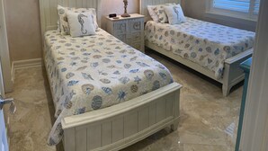Un dormitorio con dos camas, cada una con una colcha de motivos de conchas marinas, una mesita de noche con lámpara y cuadros en la pared.