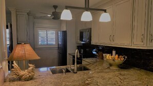 Una cocina con armarios blancos, encimera de granito, ventilador de techo y un tazón de fruta.