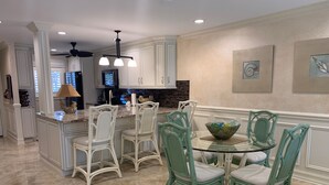 Una cocina con muebles blancos, una isla central y un comedor con sillas verdes y una mesa de vidrio.