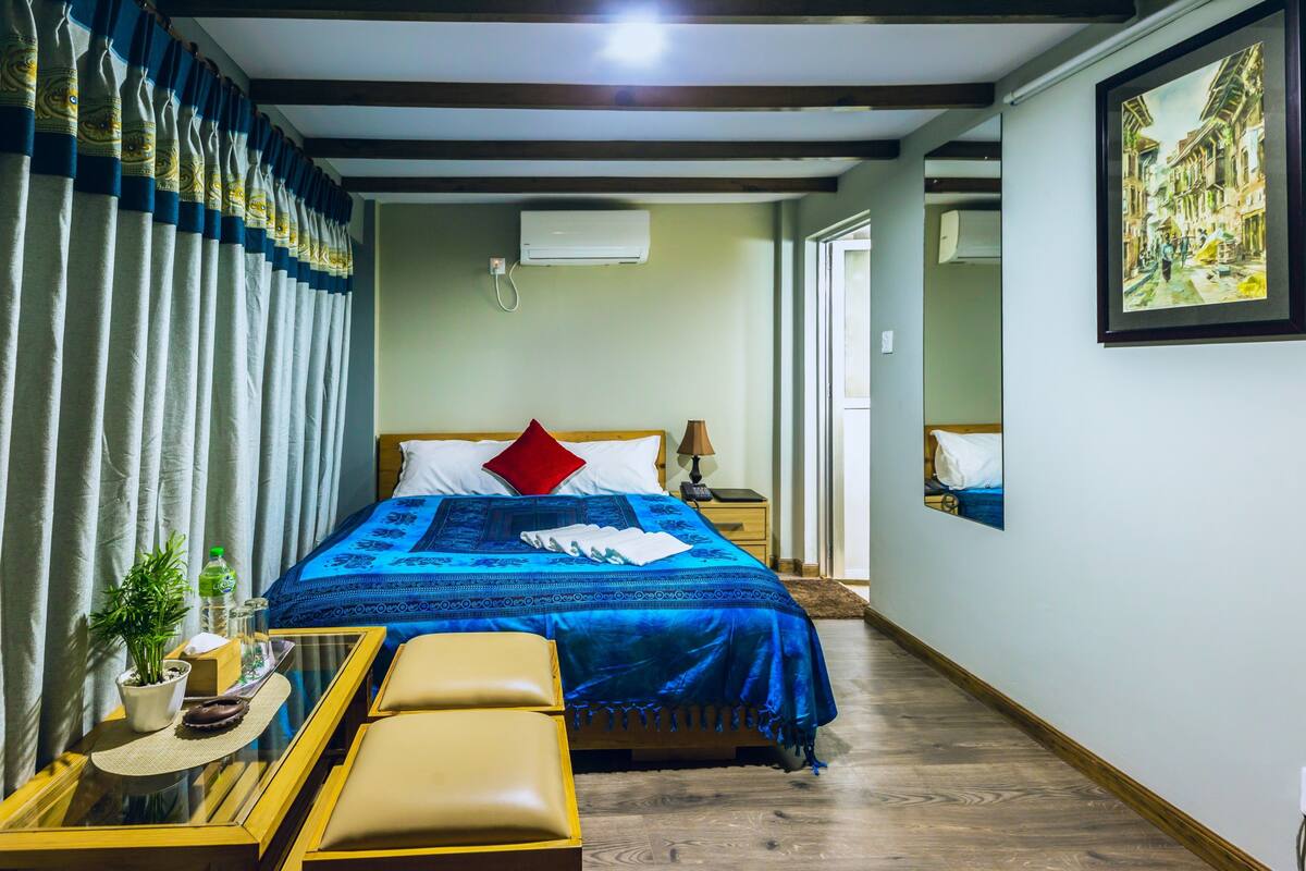 A hotel room with a bed, a bedside table with a lamp, a chair, a potted plant, and a painting on the wall.