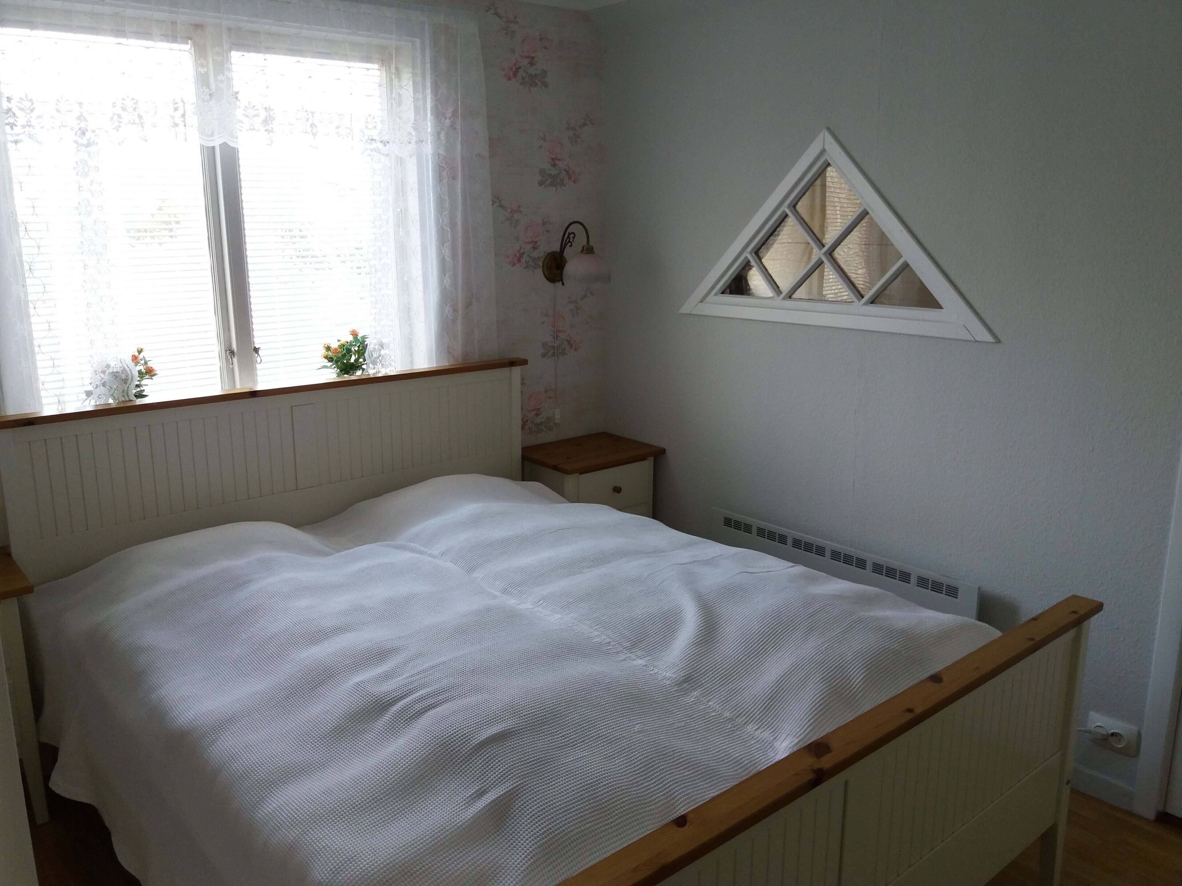 Un dormitorio con estructura de cama de madera, ropa de cama blanca, una mesita de noche pequeña, una lámpara de pared y una ventana con marco blanco.