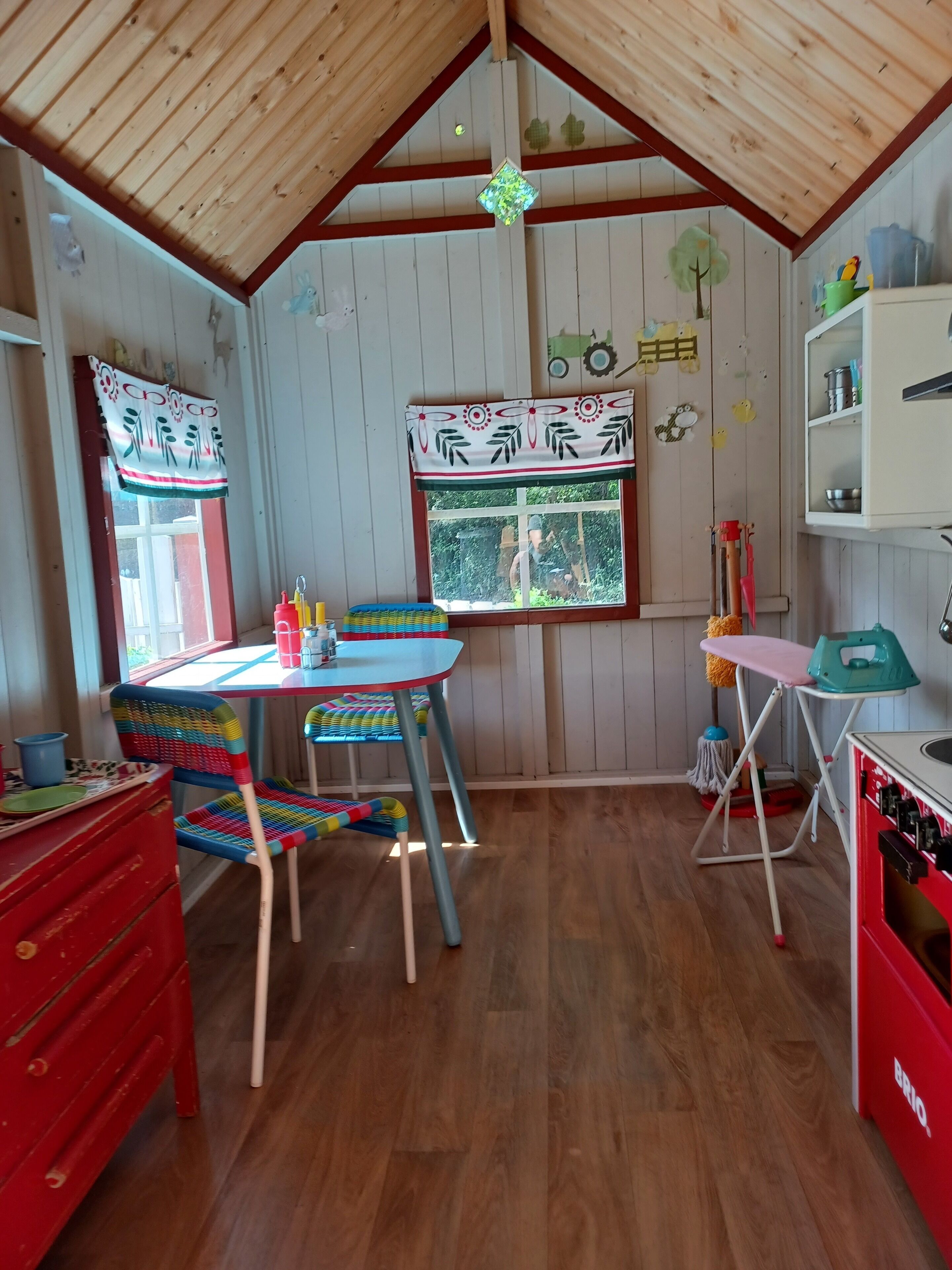 A cozy room with wooden walls, a wooden ceiling, a window with a view of greenery, a table with chairs, a red cabinet, and a shelf with items.
